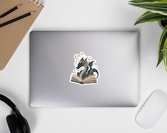 Könnte beinhalten: Ein Aufkleber mit einem blauen und goldenen Drachen, der auf einem offenen Buch sitzt, mit Sternen und einem Mond. Der Drache hat komplizierte Muster und befindet sich auf einem weißen Hintergrund. Der Aufkleber befindet sich auf einem Laptop.
