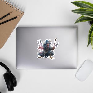 May include: A sticker depicting a fantastical creature with a chicken-like head, adorned in a blue hat and coat, wielding forks and knives. The sticker is on a laptop, with a notebook, headphones, and a mouse in the background.