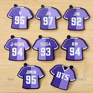 May include: A collection of purple and white jersey-shaped keychains. Each keychain features a player's name or initials and a number. One keychain displays "BTS" on the front. The keychains are arranged on a wooden surface.