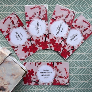 Peut inclure: Enveloppes à motifs floraux avec des fleurs rouges et des accents blancs. Chaque enveloppe présente une étiquette circulaire avec du texte français, dont "Maison", "Shopping", "Pharmacie" et "Courses". Un portefeuille est visible.