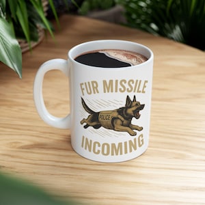 Könnte beinhalten: Weiße Keramik-Kaffeetasse mit schwarzem Kaffee. Die Tasse zeigt eine Grafik eines laufenden Schäferhundes mit Polizei-Weste. Der Text auf der Tasse lautet "FUR MISSILE INCOMING" in einem beigen Farbton.