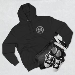 May include: Black hooded sweatshirt featuring a white Maltese cross emblem with the text "FIRST IN LAST OUT". The image also shows dark wash jeans, a white belt, and black and white trainers.