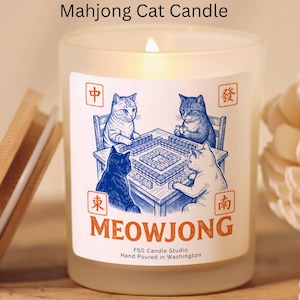 May include: Frosted glass candle with a lit wick. The label features a whimsical illustration of four cats playing mahjong. The word "MEOWJONG" is printed in orange, with the text "FSG Candle Studio" and "Hand Poured in Washington" below.