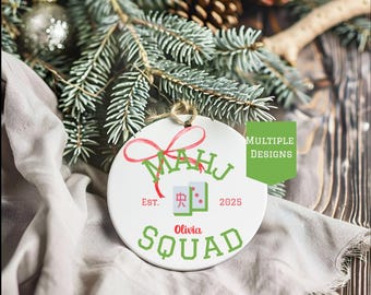 May include: A white ceramic ornament with a red bow and green text that reads "MAHJ" and "Olivia Squad". The ornament also features a graphic of two Mahjong tiles and the year 2025. The ornament is hanging from a twine loop.