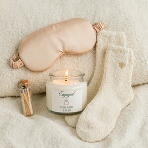 May include: A candle labeled "Engaged" with a lit wick, a satin eye mask, a pair of fuzzy socks with a gold heart, and a small glass bottle of matches. The items are arranged on a soft, white surface.
