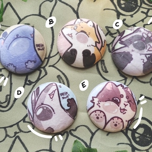 Pode incluir: Uma coleção de cinco botões redondos com ilustrações de animais de desenho animado. Os botões apresentam um elefante azul, uma raposa, um guaxinim, um texugo e um gato. Cada botão tem um design e esquema de cores únicos, com o texto "Belle Skull".