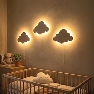 Könnte beinhalten: Drei wolkenförmige Wandleuchten mit warmer Hintergrundbeleuchtung und silbernen Zugketten, montiert an einer Wand über einem Holzbettchen. Im Bettchen liegt ein wolkenförmiges Plüschkissen mit gestickten geschlossenen Augen und einem Lächeln.