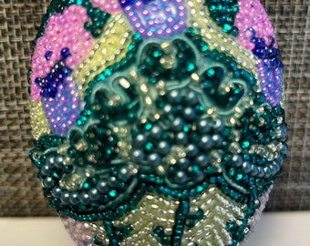 May include: A beaded Easter egg with a floral design. The egg is decorated with pink, purple, blue, and green beads. The egg is sitting on a clear stand.