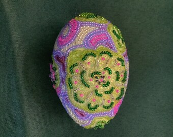 May include: A decorative egg with a green, purple and pink beaded design. The egg is covered in small beads that create a floral pattern.