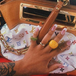 May include: A brown leather handbag with gold hardware and a white patterned interior. The bag is next to a hand with several rings, including a green and yellow ring. The nails are painted with a white tip.
