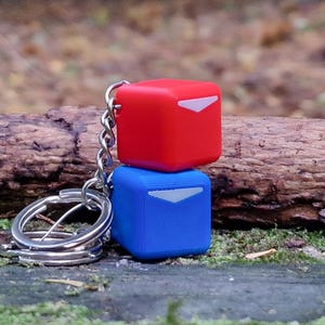 May include: A red and blue cube-shaped keychain. The red cube sits atop the blue cube, both with a white triangular design. The keychain is attached to a silver chain and ring. The background features a log and moss.