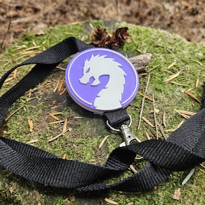 Puede incluir: Un cordón negro con un cierre plateado y una insignia redonda, morada y blanca con un diseño de dragón estilizado. El cordón descansa sobre una superficie verde musgosa con agujas de pino y una piña pequeña.