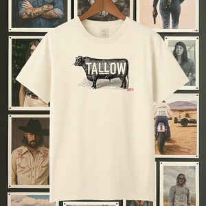 May include: Cream-colored t-shirt with a black illustration of a bull and the word "TALLOW" in bold, white letters. The shirt is hanging on a wooden hanger, surrounded by framed photographs with a rustic theme.