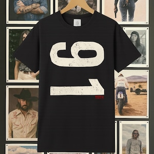 Może przedstawiać: Czarny t-shirt z białymi cyframi "91" i czerwonym napisem "TUTS". Koszulka wisi na drewnianym wieszaku na tle ściany ozdobionej oprawionymi zdjęciami w stylu vintage.