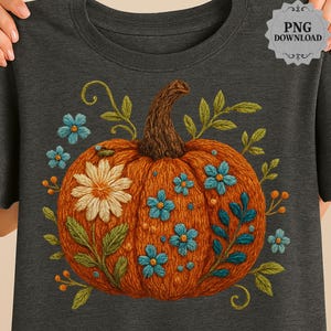 Puede incluir: Camiseta gris oscuro con una calabaza naranja bordada, un tallo marrón, flores blancas y azules y hojas verdes. La camiseta tiene cuello redondo y mangas cortas. La imagen incluye un gráfico "PNG DOWNLOAD".