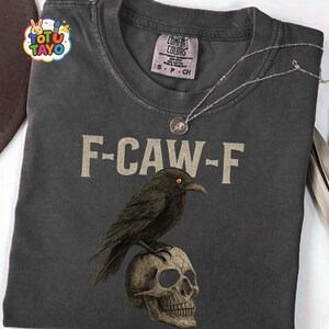 May include: Dark gray t-shirt with the text "F-CAW-F" and a graphic of a black crow perched on a skull. A silver necklace with a round pendant rests on the shirt. The shirt has a Comfort Colors tag.
