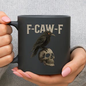 May include: A black ceramic mug featuring a raven perched atop a skull. The mug has the text "F-CAW-F" in a vintage font. The raven has orange eyes. The mug is held by a person.