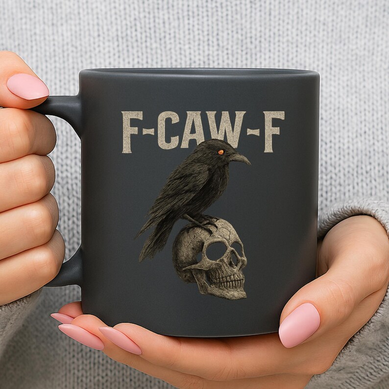 May include: A black ceramic mug featuring a raven perched atop a skull. The mug has the text "F-CAW-F" in a vintage font. The raven has orange eyes. The mug is held by a person.