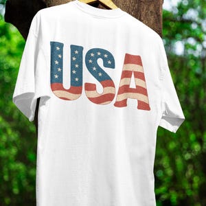 Puede incluir: Camiseta blanca con la palabra "USA" impresa en la espalda. Las letras están diseñadas con la bandera estadounidense, con azul con estrellas blancas y rayas rojas y blancas. La camiseta está colgada en una percha de madera.