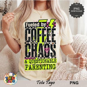 May include: A light beige t-shirt featuring the text "Fueled by Coffee Chaos & Questionable Parenting" in black, neon green, and pink. The design includes lightning bolts and a heart. A clear glass is held in the hand.