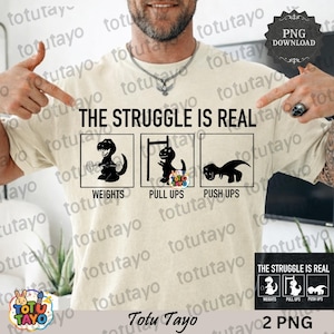 May include: A beige t-shirt with the text "THE STRUGGLE IS REAL" and three black dinosaur illustrations representing weightlifting, pull-ups, and push-ups. The shirt has a crew neck and short sleeves.