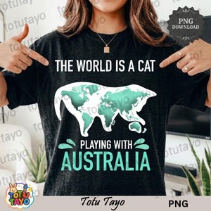 May include: Charcoal grey t-shirt featuring the phrase "THE WORLD IS A CAT" above a cat-shaped world map in white and teal. The text "PLAYING WITH AUSTRALIA" is below in teal. The shirt has a relaxed fit.