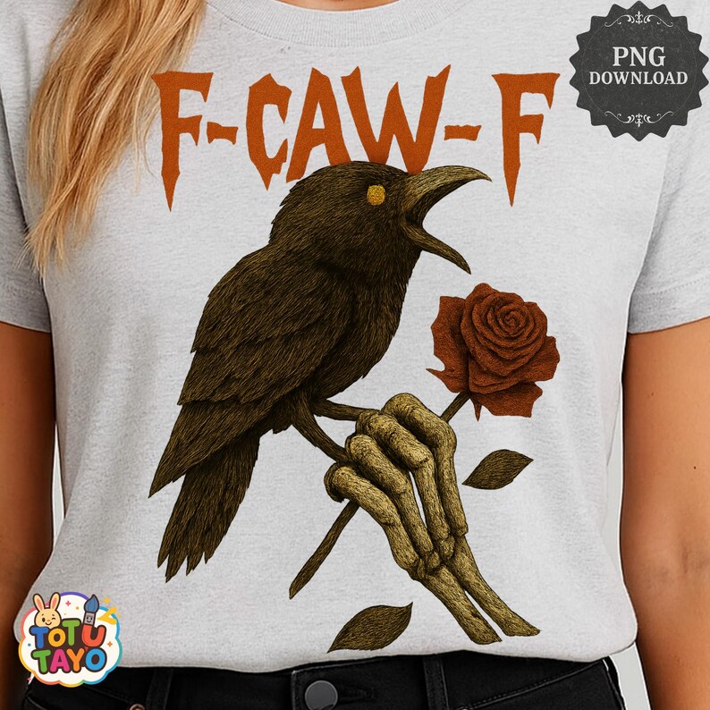 May include: A light gray t-shirt featuring a detailed illustration of a crow perched on a branch, holding a rose. The crow has yellow eyes and is set against the text "F-CAW-F" in orange. The design includes a skeleton hand holding the rose.