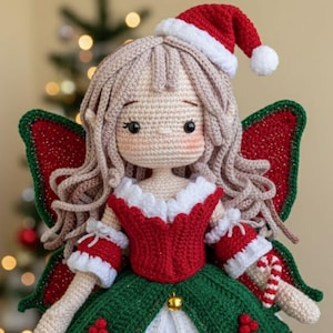 Könnte beinhalten: Eine handgefertigte gehäkelte Weihnachtsfeenpuppe. Die Puppe hat ein rotes und grünes Kleid, Flügel und einen Weihnachtsmannhut. Die Puppe hat blondes Haar und rosige Wangen. Die Puppe ist mit goldenen Glocken und roten Beeren verziert. Die Puppe ist etwa 25 cm groß.