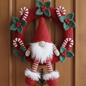 Könnte beinhalten: Ein handgefertigter Weihnachtskranz mit roter Häkelbasis, verziert mit grünen Stechpalmenblättern, roten Beeren und Zuckerstangen-Akzenten. Ein Wichtel mit roter Mütze, weißem Bart und gestreiften Beinen ist das Herzstück.