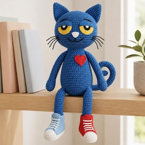 May include: A blue crocheted cat doll with yellow eyes, a white nose, and black whiskers. The cat features a red heart on its chest and wears one light blue sneaker and one red sneaker with white laces. The doll is sitting upright with a curled tail.