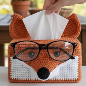 Puede incluir: Una funda de caja de pañuelos de ganchillo naranja hecha a mano con forma de zorro, completa con orejas, hocico blanco y nariz negra. Gafas con montura negra descansan sobre la cara del zorro. Una mano saca un pañuelo de la abertura.