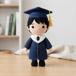 Puede incluir: Muñeca de ganchillo hecha a mano vestida con una toga y birrete de graduación. La muñeca lleva una toga azul marino con cuello blanco y un gorro a juego con borla dorada. La muñeca tiene el pelo negro y está de pie.