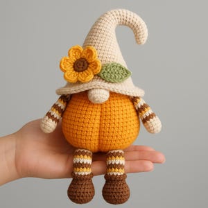 Puede incluir: Muñeca de gnomo de ganchillo hecha a mano con cuerpo en forma de calabaza. El gnomo lleva un sombrero color crema adornado con un girasol y una hoja verde. Sus brazos y piernas tienen patrones de rayas en marrón y crema. Decoración temática de otoño.