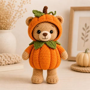 Puede incluir: Un oso de peluche de ganchillo color canela con un disfraz de calabaza naranja brillante. El disfraz cuenta con una capucha con un tallo marrón y una hoja verde, un cuello de hojas verdes y un cuerpo de calabaza acanalado. El oso tiene ojos negros.