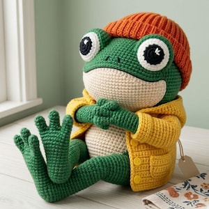 Op de afbeelding: Een handgemaakte gehaakte kikkerpop met grote, expressieve ogen, een oranje muts en een gele jas. De kikker is groen met een crèmekleurige buik en zit met uitgestrekte benen. Een charmant, grillig pluche speelgoed.