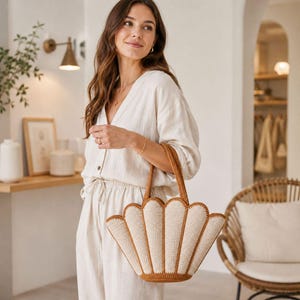 Peut inclure: Un sac fourre-tout en forme de coquillage, de couleur crème, avec des bordures et des poignées marron. Le sac est fait d'un matériau crocheté et est tenu par une personne vêtue d'une tenue crème.
