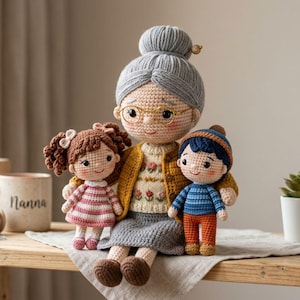 Op de afbeelding: Een handgemaakte gehaakte poppenset met een omafiguur met een bril, een gele cardigan en een grijze knot, geflankeerd door een meisje in een gestreepte jurk en een jongen in een blauw en oranje outfit. De poppen zitten op een houten oppervlak.