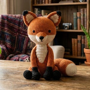 以下が含まれることがあります： オレンジ、白、黒のディテールが施された手作りのかぎ針編みのキツネ人形。キツネは尖った鼻、大きな耳、ふわふわの尻尾を持っています。木製の表面に座っています。背景には本棚と模様入りの毛布が含まれています。