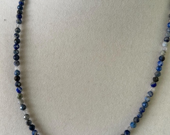 May include: A beaded necklace featuring small, faceted beads in shades of blue, gray, and white. The necklace is strung on a clear cord and is displayed on a white mannequin.