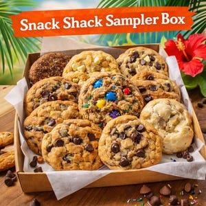 May include: A variety of freshly baked cookies in a cardboard box. The cookies include chocolate chip, white chocolate chip, and cookies with colorful candies. The box is labeled "Snack Shack Sampler Box". A red hibiscus flower is visible in the background.