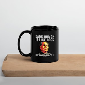 Puede incluir: Taza de cerámica negra con el texto "DARK HUMOR IS LIKE FOOD NOT EVERYONE GETS IT" y un retrato de un hombre. La taza está sobre una superficie de madera.