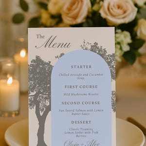 Pode incluir: Um cartão de menu de casamento com uma seção arqueada azul clara listando os pratos: Sopa fria de abacate e pepino, Risoto de cogumelos silvestres, Salmão grelhado e Tiramisu clássico. O menu está em um prato branco.