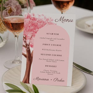 Pode incluir: Um cartão de menu de casamento com um design floral rosa e a palavra "Menu" no topo. O menu lista uma entrada, um primeiro prato, um segundo prato e uma sobremesa. Os nomes "Mariana + Pedro" estão na parte inferior.