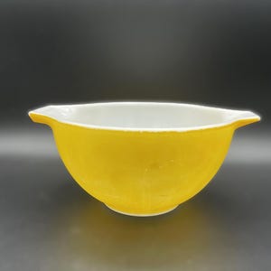 May include: A vintage yellow mixing bowl with a white interior. The bowl has a rounded shape with a flared rim and two small handles. The bowl is made of a durable material and is suitable for kitchen use.
