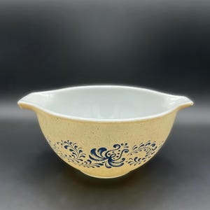 May include: A vintage mixing bowl with a speckled tan exterior and a white interior. The bowl features a blue floral design around the exterior. It has two handles on either side.