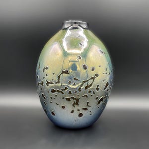 May include: An iridescent, oval-shaped vase with a textured surface. The vase transitions from a light green at the top to a blue-silver at the bottom, with dark brown, irregular patterns. The vase has a small, dark rim at the top.