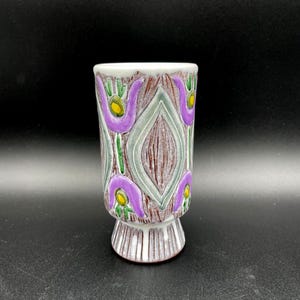 May include: A ceramic vase with a white interior and a decorative exterior. The exterior features a pattern of purple tulip-like shapes, green accents, and yellow centers on a brown background. The vase has a flared base and a cylindrical body.