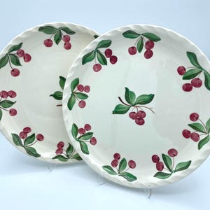 May include: Two white ceramic plates with a scalloped edge. Each plate is decorated with a repeating pattern of red cherries and green leaves. The plates are displayed on clear stands against a white background.