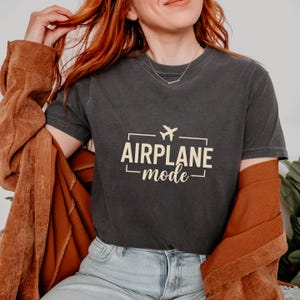 May include: Dark gray t-shirt with the text "AIRPLANE mode" in a rectangular frame, featuring a small airplane icon. The shirt is worn with a brown jacket.