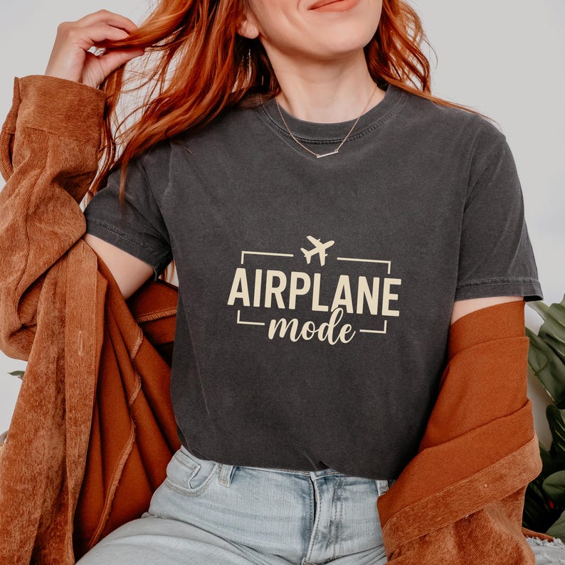 May include: Dark gray t-shirt with the text "AIRPLANE mode" in a rectangular frame, featuring a small airplane icon. The shirt is worn with a brown jacket.